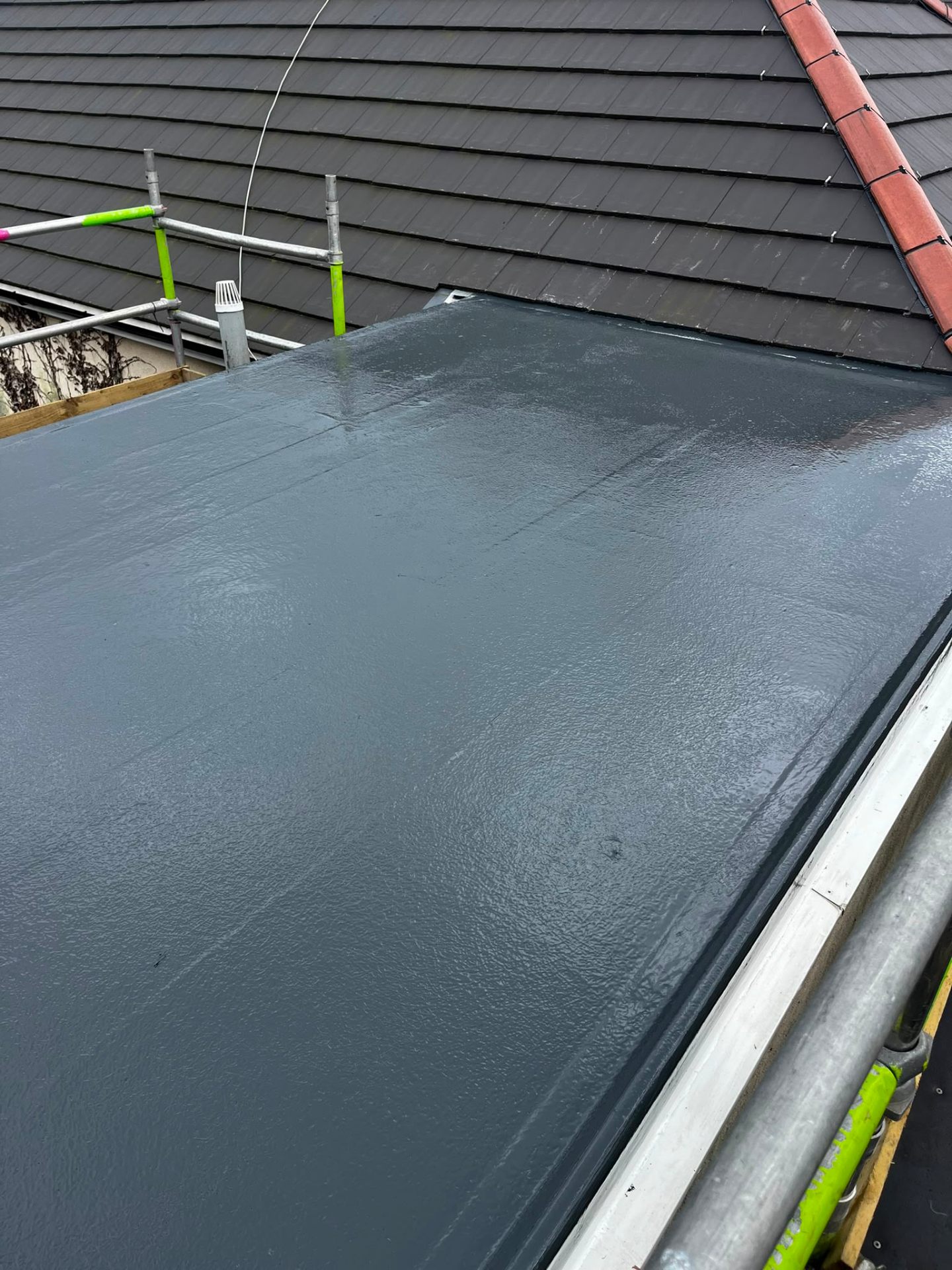 Freshly laid dark-grey GRP fibreglass flat roof with clean upstand fillet against slate tile abutment, scaffolded and completed by RTC Roofing on the Wirral