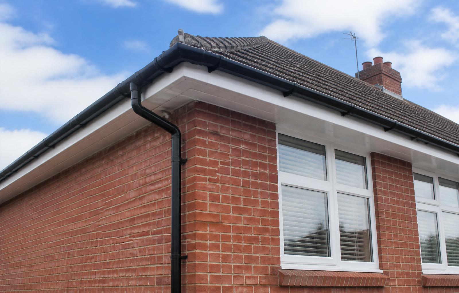 Newly installed black uPVC half-round guttering and downpipe on a red brick British semi with white fascia and soffit boards
